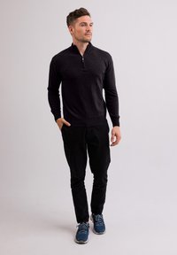 Tmavě šedý zip-up svetřík s vysokým límcem, v kombinaci s černými kalhotami a modrými sportovními botami. Svetřík má těsný střih a žebrované manžety.