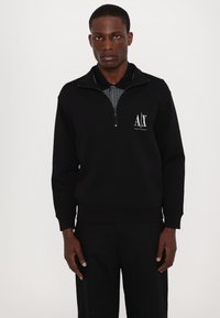 Felpa nera con zip, caratterizzata da un colletto alto, polsini a coste e un logo bianco sul petto. Progettata con una texture di tessuto morbido.