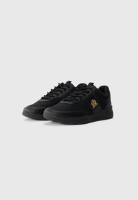 Black sneakers with a smooth synthetic upper, round toe, lace-up design, and a gold logo accent on the side. Thick rubber sole.