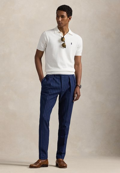 Kreminės spalvos megztas polo marškinėlis, tamsiai mėlyni dryžuoti kostiuminės kelnės, rudi odiniai batai, saulės akiniai kabančiai ant apykaklės, aptempianti siluetas.