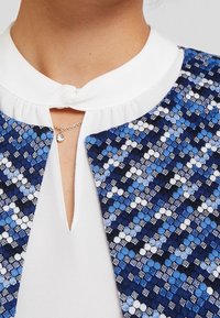 Niebiesko-biała kurtka w wzory z białym kołnierzem i dekoltem w serek, charakteryzująca się misterną teksturą tkaniny i metalowym wisiorkiem.