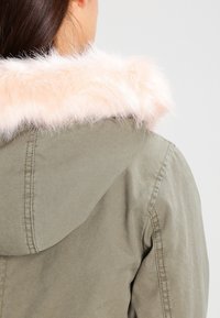Žena v olivově zelené bundě s kapucí lemovanou světle růžovou umělou kožešinou, zobrazená zezadu a mírně otočená doprava.