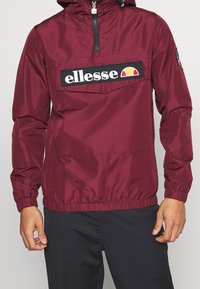 Maroon windbreaker jacket with a black logo patch, front zipper, drawstring hood, and elastic cuffs. Lightweight, smooth texture.