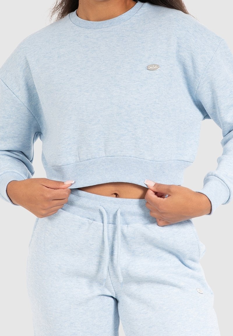 Personne portant un sweat-shirt court bleu clair et un pantalon de jogging assorti, tenant l'ourlet du sweat-shirt pour montrer un ventre nu.