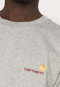 Szary bawełniany t-shirt z okrągłym dekoltem, z żółto-bordowym haftowanym logo na piersi. Miękka faktura, luźny krój.