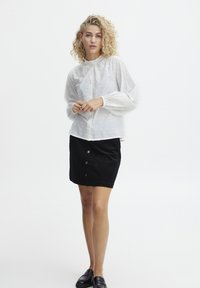 Weiße strukturierte Bluse mit Puffärmeln, kombiniert mit einem schwarzen Cordrock mit vorderen Knöpfen. Modell trägt schwarze Schuhe.