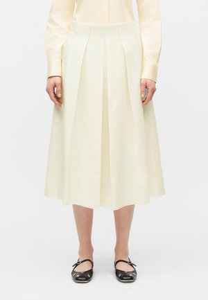 Žena oblečená v plisovanej krémovej midi sukni, bledozltej dlhorukávovej blúzke a čiernych topánkach Mary Jane s kovovými ozdobami.