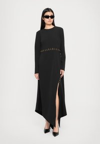 Black long-sleeve dress featuring a high slit, gold accent detailing at the waist, and a textured fabric with an uneven hemline.