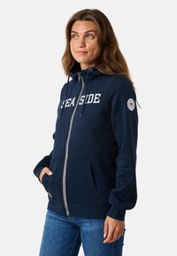 Veste à capuche zippée marine avec "SEASIDE" imprimé en blanc, capuche à cordons, poches avant et patch logo circulaire sur la manche gauche.