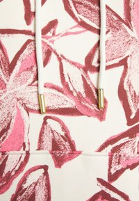 Textil con estampado floral que presenta hojas rosadas y burdeos sobre un fondo crema, con cordones blancos y detalles metálicos dorados.