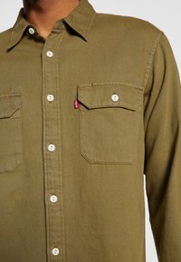 Oliwkowa koszula zapinana na guziki wykonana z bawełny, z dwoma kieszeniami na piersi, białymi guzikami i małą czerwoną metką z logo. Tekstura tkaniny o wyraźnej strukturze.