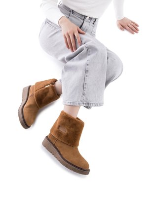 Botas de ante marrón a la altura del tobillo con un puño de piel de lana, suela de goma y costuras decorativas. El diseño incluye una cremallera lateral para ponerlas y quitarlas fácilmente.