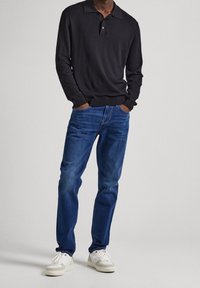 Svart långärmad pikétröja med krage, kombinerad med blå denimjeans och vita sneakers, med en avslappnad passform och klassisk design.