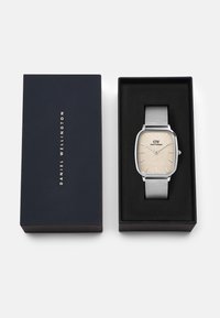 Orologio con cinturino in rete argentata, quadrante rettangolare beige, indici delle ore argentati e logo del marchio in evidenza. Confezionato in una scatola nera.