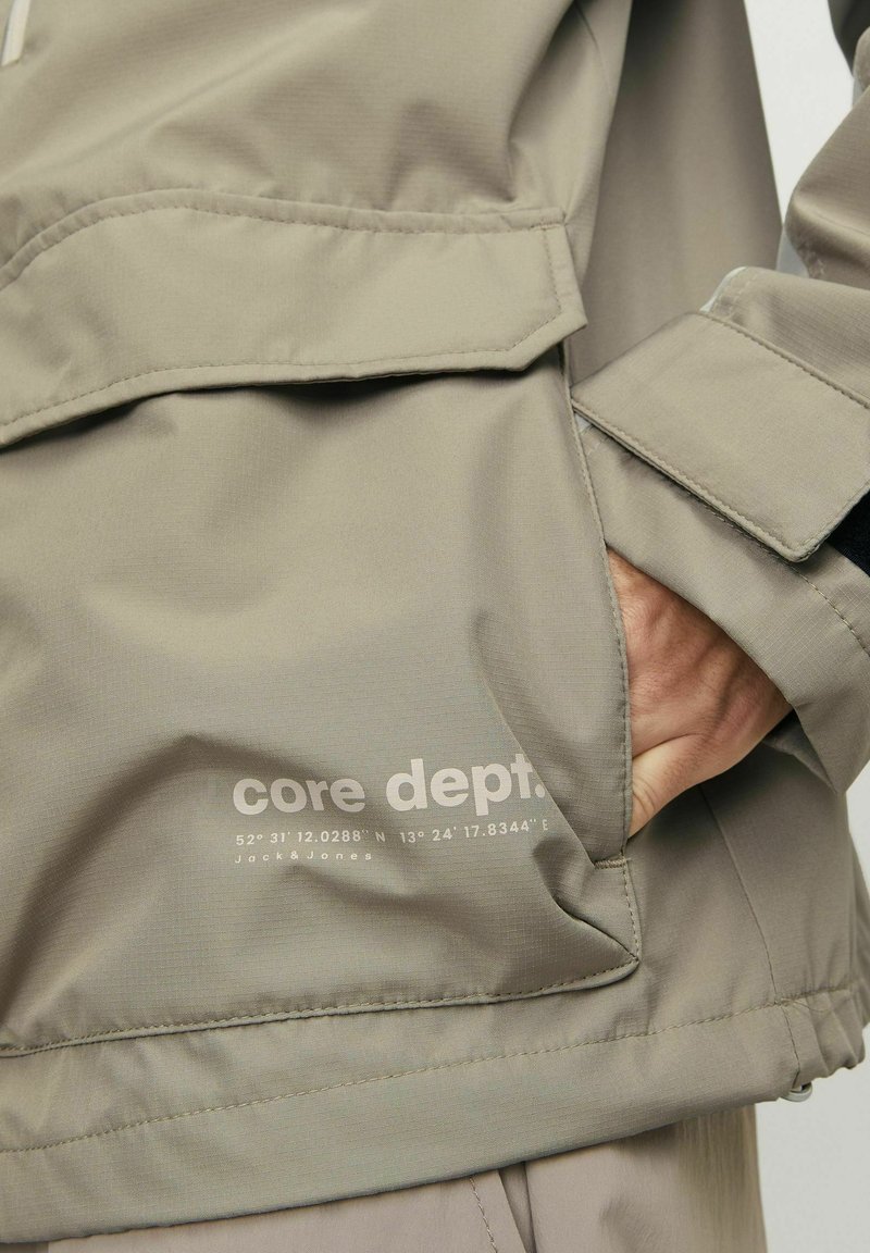 Lichtbeige jas van een gladde, gestructureerde stof. Voorzien van een voorkantzak, verstelbare manchetten en bedrukte branding aan de onderzijde.