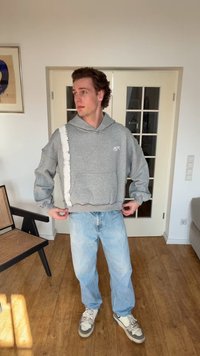 Mladić u sivoj dukserici s kapuljačom i svijetloplavim trapericama stoji u zatvorenom prostoru te podešava donji rub dukserice rukama.