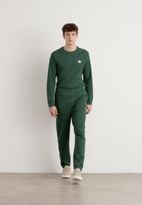 Grønn lounge-sett laget av mykt stoff, med genser med rund hals og lange ermer, samt bukser med elastisk liv. Modellen har på seg beige sneakers.
