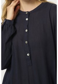 Femme portant un chemisier bleu marine à manches longues avec des boutons blancs sur le devant et des poignets boutonnés, touchant son cou d'une main.