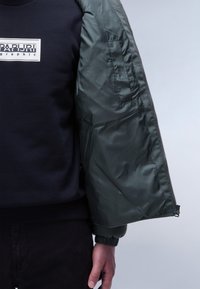 Giacca a vento trapuntata con una finitura lucida verde scuro, dotata di chiusura zip e polsini a coste, indossata sopra una felpa blu navy con un logo grafico.