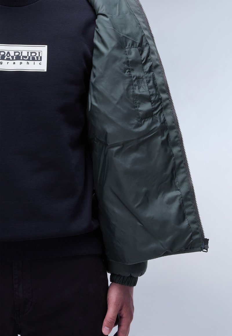 Dicker daunenjacke mit einem dunkelgrünen, glänzenden Finish, ausgestattet mit einem Reißverschluss und gerippten Bündchen, getragen über einem marineblauen Sweatshirt mit einem grafischen Logo.