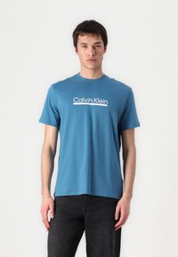 Tyrkysové bavlnené tričko s okrúhlym výstrihom a krátkymi rukávmi, s bielym nápisom "Calvin Klein" potlačeným na hrudi.