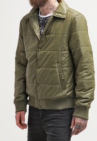 Olivgrüne Pufferjacke mit Kragen, Druckknopfverschluss, gerippten Bündchen und seitlichen Taschen. Hat eine gesteppten Textur.