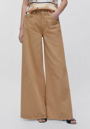 Persona che indossa pantaloni beige a vita alta e gamba larga con tasche frontali, abbinati a un top di seta con motivo floreale e tacchi neri aperti in punta.