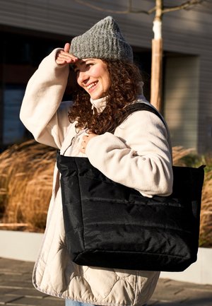 Jonge vrouw in winterjas en grijze gebreide muts draagt een grote zwarte totebag, glimlacht en houdt haar hand boven haar ogen om zich tegen de zon te beschermen in een zonnige buitenomgeving.
