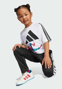 Biała koszulka krótki rękaw z czarnym logo Adidasa i grafiką Spider-Mana, w zestawie z czarnymi spodniami i białymi butami z czerwono-niebieskimi akcentami.
