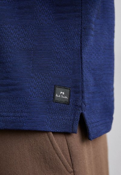 Marinefarget teksturert skjorte med et geometrisk mønster, med en svart etikett med hvit tekst. Brune bukser delvis synlige nedenfor.