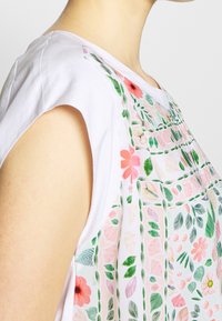 Mujer con una blusa sin mangas blanca decorada con bordados florales en rosa y verde en el frente y el hombro, contra un fondo liso.