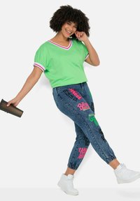Camiseta verde de cuello en V con rayas contrastantes en rosa y blanco en las mangas, combinada con joggers de mezclilla azules con gráficos coloridos. Zapatillas blancas.