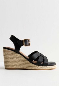 Black leather wedge sandal with a woven jute platform, double strap design, and a gold buckle accent. Smooth texture and rounded toe.