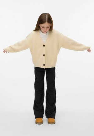 Beige strik cardigan med v-hals og tre store brune knapper, parret med sorte brede bukser og lysebrune sko.