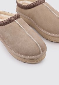 Zapatillas de casa de ante beis sin cordones, con un cuello de punto marrón texturizado. Presentan costuras blancas y una suela de goma con detalles acanalados.