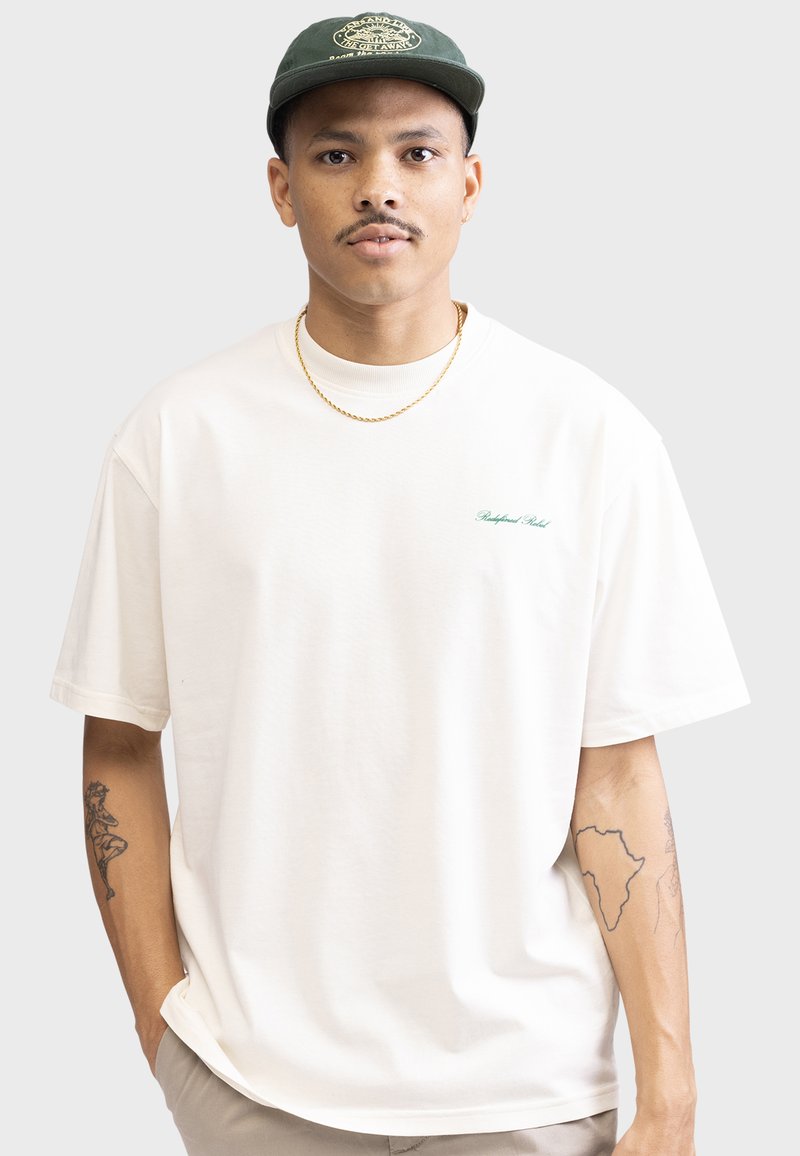 T-shirt en coton de couleur crème, ample, avec col rond côtelé, manches courtes et petit logo vert sur la poitrine.