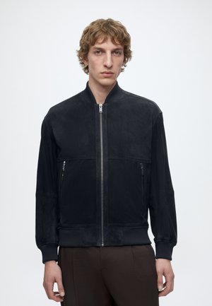 Jonge man met krullend blond haar, die een zwarte bomberjas met rits en een bruine broek draagt, staand tegen een effen achtergrond.