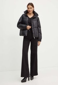 Giacca a vento nera con colletto alto, tessuto lucido, polsini elasticizzati e chiusura a zip. Abbinata a pantaloni neri a vita alta e con fondo svasato, e tacchi a piattaforma.