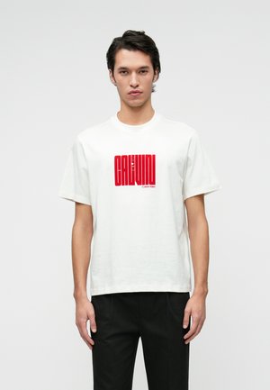 Bílý bavlněný tričko s krátkým rukávem, které obsahuje červený grafický potisk se textem "CALVIN KLEIN." Volný střih, kulatý výstřih.
