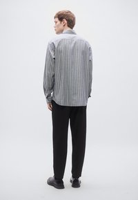 Szürke-fehér csíkos ing, hosszú ujjú, gombos gallérral. Fekete nadrág és sötét cipő. Álló póz. Minimalista háttér.