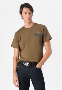 Oliven grønn bomull t-skjorte med korte ermer, med et svart grafisk merke på venstre bryst. Sammen med mørkeblå jeans og et svart belte.