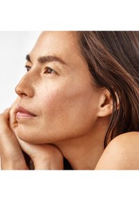 Femme aux longs cheveux bruns reposant son visage sur ses mains, regardant pensivement sur le côté contre un fond blanc uni.