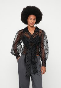 Blouse transparente noire à pois avec un design croisé, manches bouffantes longues, col et un détail de taille nouée, assortie à un pantalon gris.