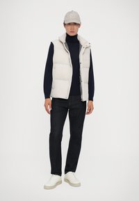 Weste in hellbeige, langärmliger marineblauer Pullover, dunkle Jeans und weiße Sneakers mit beigen Sohlen; Baseballmütze mit Logo.