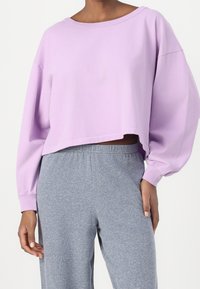 Vaalee purppuravärinen lyhyt huppari, jossa on pitkät hihat, kevyt kangas ja leveä pääntie, yhdistettynä harmaisiin joustaviin vyötäröhousuihin.