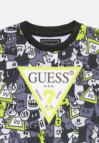 T-shirt graphique avec un imprimé cartoon coloré sur fond noir, avec un logo triangulaire jaune proéminent étiqueté "GUESS U.S.A."