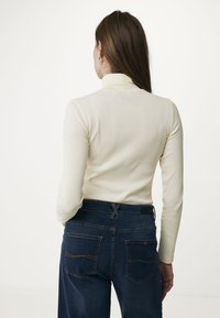 Cremfarbener Rippstrick-Longsleeve mit Stehkragen, kombiniert mit dunkelblauen, hoch taillierten Jeans, die zwei Gesäßtaschen und kontrastierende Nähte aufweisen.