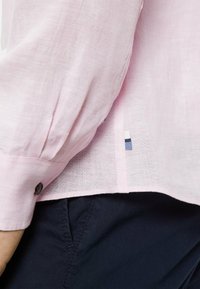  Lichtroze linnen shirt met opgerolde mouwen, met een knoopdetail. De zijkant heeft een contrasterend blauw-wit label. Gecombineerd met donkere broeken.