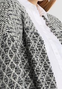 Mujer vistiendo un cárdigan de punto texturizado en blanco y negro sobre una camisa blanca de botones, mostrando la parte superior del torso y el área del collar.