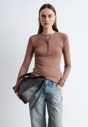 Femme portant un haut marron ajusté à manches longues et un jean bleu clair, tenant un sac à main en cuir noir, une main dans la poche.